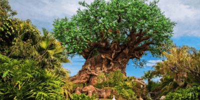 A large, intricate artificial tree with bright green leaves and detailed carvings on its trunk stands amid lush tropical plants under a blue sky—a true icon for any fan of Best Disney World Trivia.