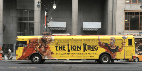Large yellow bus photographed in the city with The Lion King Musical promotion signage displayed across the bus