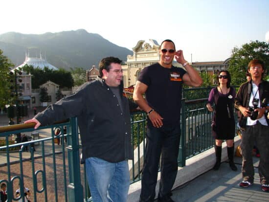 Dwayne Johnson photographed smiling with left arm near his ear, while wearing an all-black outfit and sunglasses