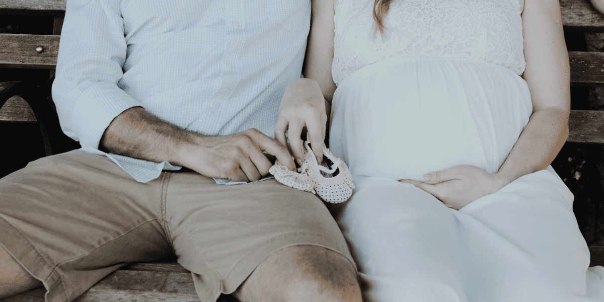 Pregnant couple holding tiny pink baby shoes while sitting on wooden bench for pregnancy announcement photo