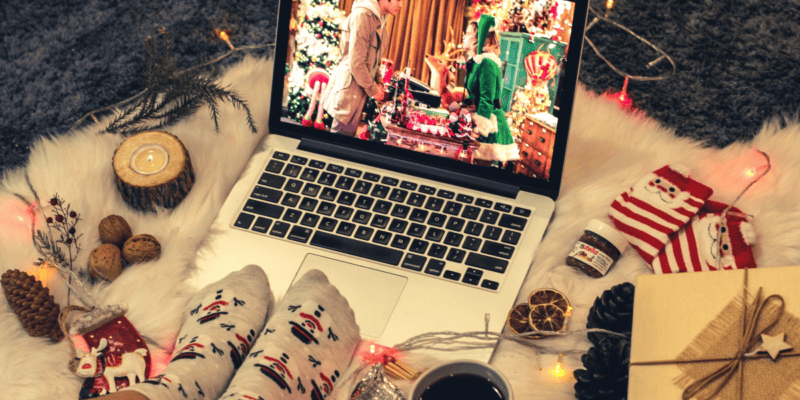A laptop displaying a Christmas movie on the floor on top of a white fur carpet, surrounded by a small jar of Nutella, Christmas socks, and other Christmas decor