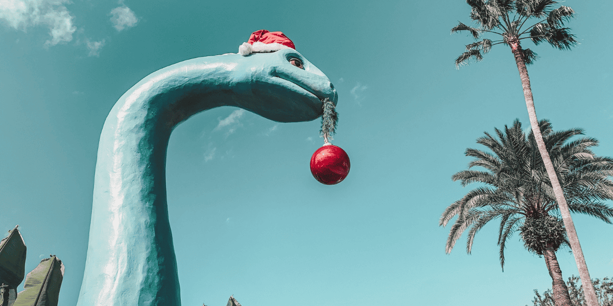 A giant blue dinosaur character at Echo Lake, Hollywood Studios pictured with a Christmas hat on its head and Christmas red ornament hanging from its mouth