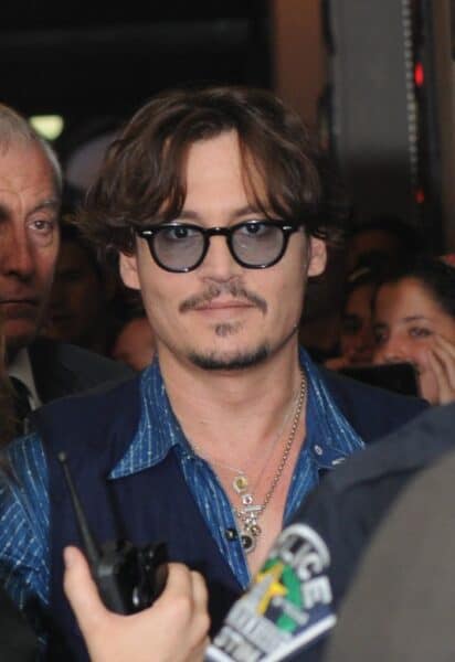 Johnny Depp looking at the camera while wearing a navy blue vest, layered with a lighter blue shirt and accessorized with several necklaces. While looking at the camera, Johnny Depp is holding a walkie-talkie device.