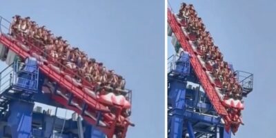 Guests aboard Six Flags’ iconic red and blue roller coaster are paused dramatically on an angled track high above the park, their expressions a mix of excitement and suspense against the backdrop of a clear sky, as whispers among thrill-seekers recall moments reminiscent of Final Destination and tales of legendary coaster mishaps.