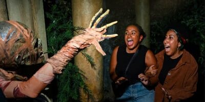 Two friends react with a scream and laughter as a costumed creature reaches out to them during Halloween Horror Nights in a spooky, dimly-lit forest setting at Halloween Horror Nights.