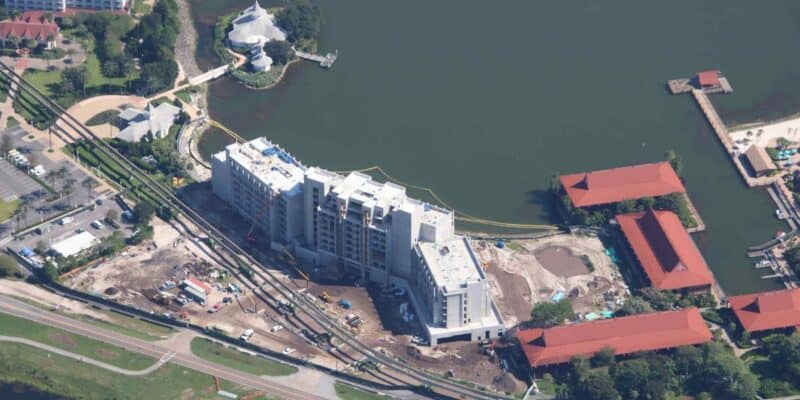 Aerial view of a construction site for a multi-story building near a lake, showcasing the site's expansion. The area is surrounded by cranes and construction vehicles. Adjacent to the site are red-roofed buildings and a white tent-like structure. Green spaces and pathways are visible.