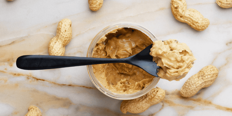 A spoon filled with peanut butter resting on top of an open jar of peanut butter, surrounded by whole peanuts.