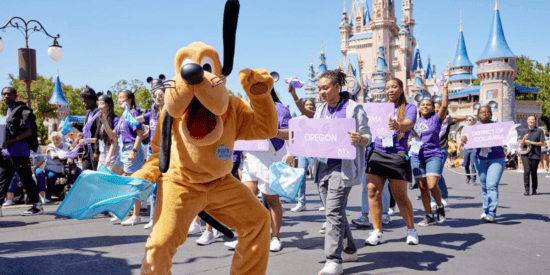 Pluto leading a procession of Disney Dreamers Academy teens displaying signs for where they come from.