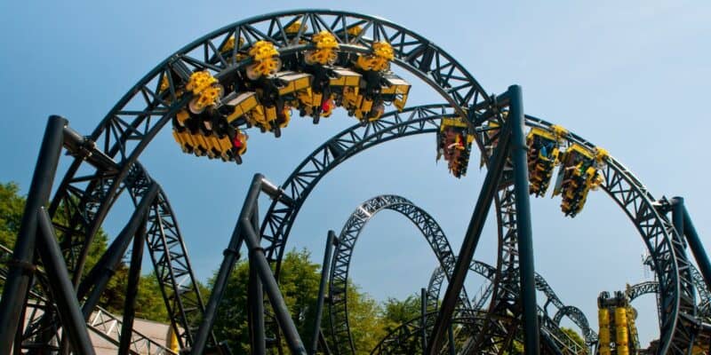 A roller coaster with an intricate series of loops and twists has passengers riding in yellow and black cars. Set against a clear blue sky with some trees visible in the background, the coaster appears to be in mid-loop, but there's no sign of any roller coaster accident marring the scene.