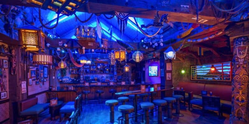 Interior of a vibrant tiki bar with colorful lighting, eclectic decor featuring nautical elements, wooden stools, and a thatched bar roof; notably one of the least kid-friendly places at Disney World.