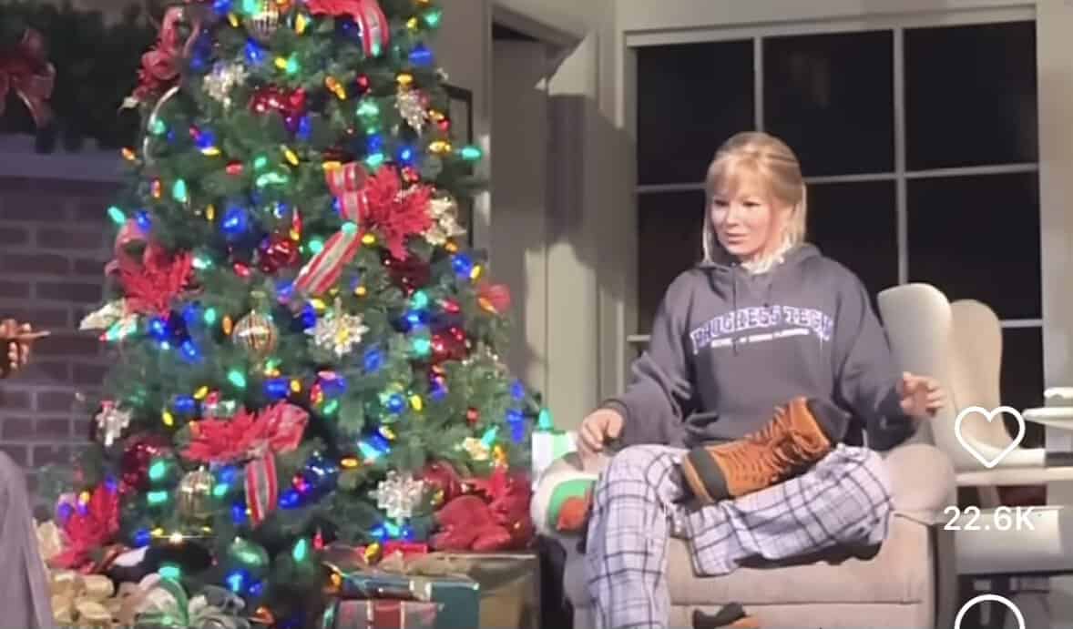 A guest dressed in festive pajamas poses by a dazzlingly decorated Christmas tree at a holiday-themed event, showing off their boots amid piles of wrapped gifts and twinkling lights—channeling the cozy, glamorous spirit of Taylor Swift’s seasonal spectacular.