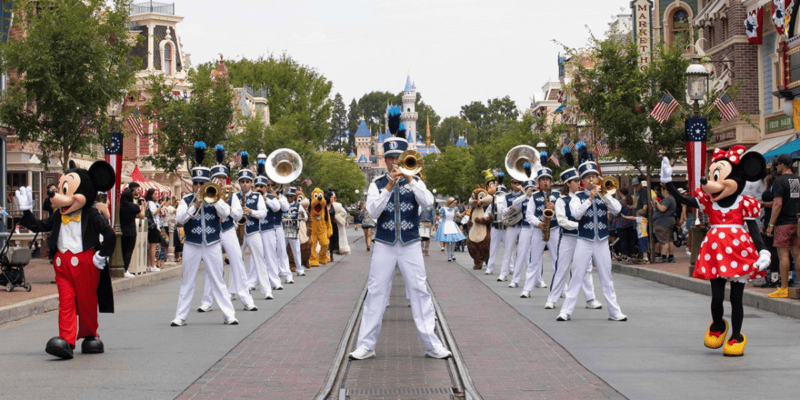 Mickey Mouse and Minnie Mouse accompanying the Disneyland Main Street Marching Band down Main Street, U.S.A, with Disneyland Park's Sleeping Beauty Castle in the background