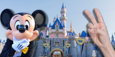Mickey Mouse shocked in front of the Disneyland Sleeping Beauty Castle with a person holding up a number two hand sign.