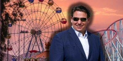 Florida Governor Ron DeSantis smiling with sunglasses on with a theme park in the background.