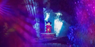 Mickey Mouse, dressed as a magician on stage during the Fantasmic! show, surrounded by dramatic blue and purple stage lights and mist during a nighttime performance at Disneyland Park.