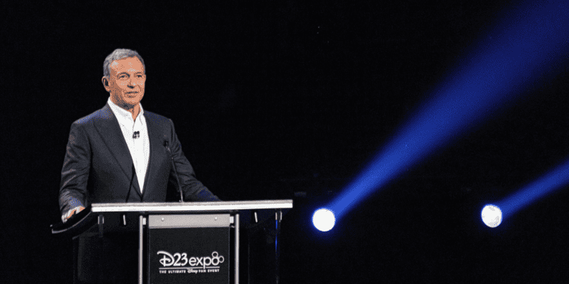 A man giving a speech at a podium with the "D23 Expo" logo, under a spotlight on a dark stage, discussing Disney’s advancements in artificial intelligence.