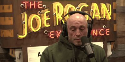 Joe Rogan looks down while talking into a podcast mic.