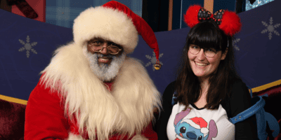 Santa Claus played by a Black Disney performer sits in a sleigh next to a woman with dark brown hair, glasses, and bangs. They're both smiling and dressed in festive gear.