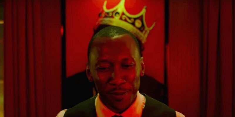 Mahershala Ali in front of a painting with a crown on it in a red room