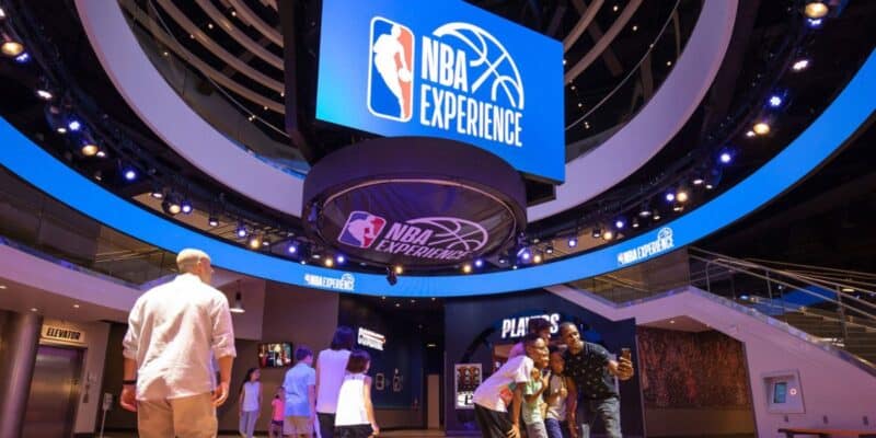 Visitors explore the NBA experience at Disney Springs, featuring interactive kiosks, basketball-themed attractions, and a large ceiling-mounted display.