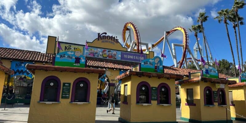 A photo of the entrance to Knott's Berry Farm. Ticket booths with vibrant signs are in the foreground. Behind them, a roller coaster's loops and tracks are visible against a partly cloudy sky, with tall palm trees scattered around the entrance area.