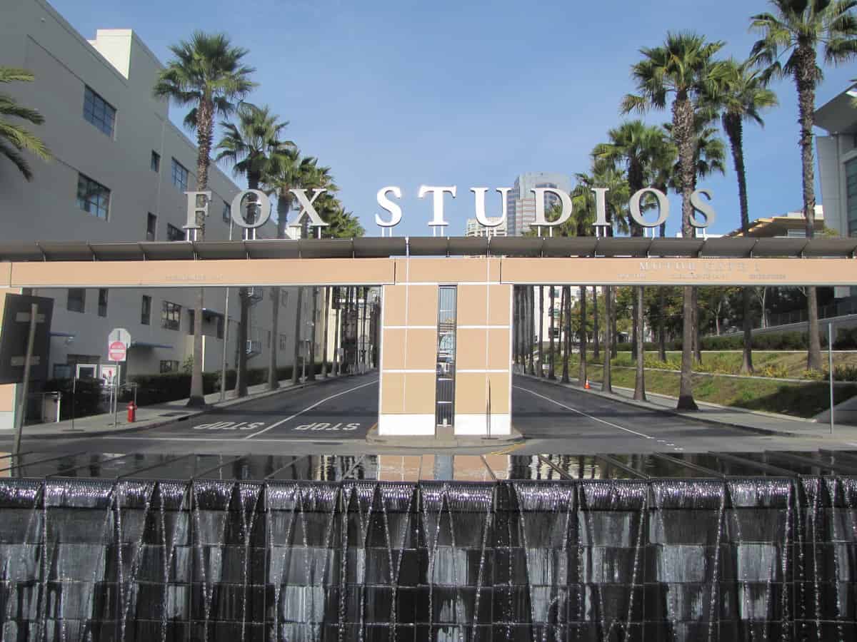 A sunny day at the entrance of fox studios, showcasing the iconic sign above the gate with a reflective water feature in the foreground.