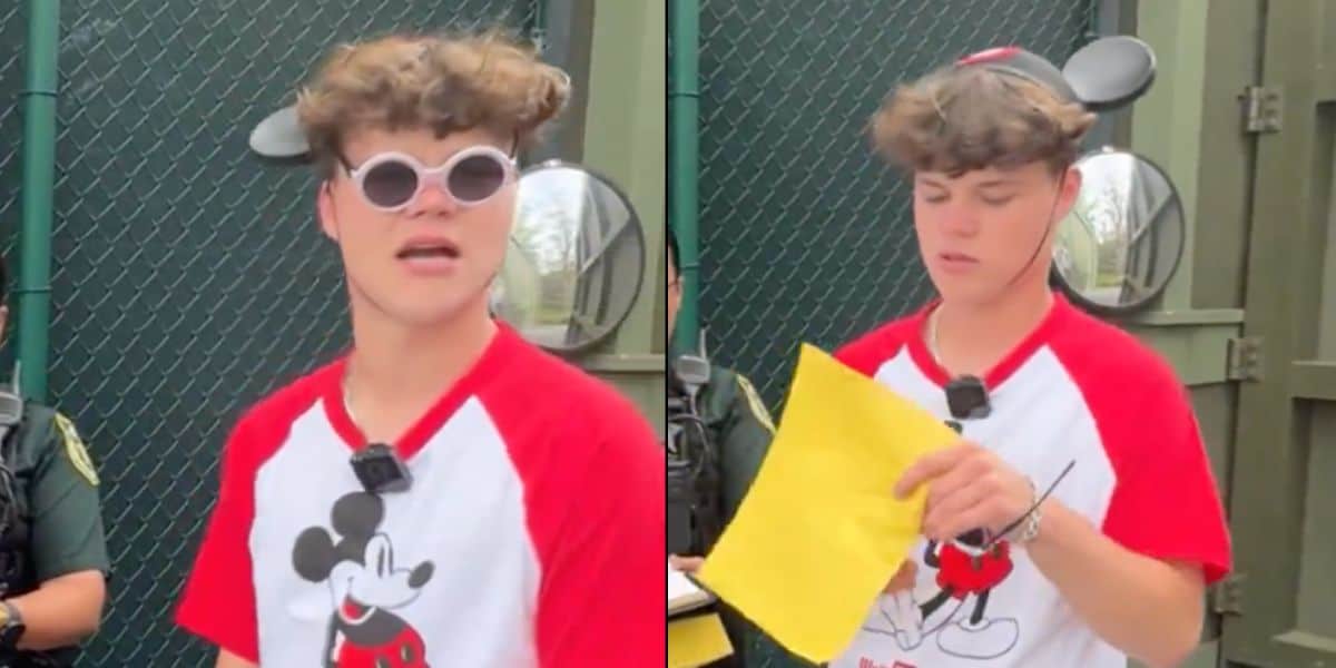 Man wearing mickey mouse t-shirt and sunglasses, holding a yellow paper, with a surprised expression outdoors near a fence.