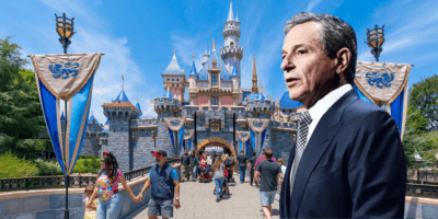A man in a suit stands in front of Disneyland’s Sleeping Beauty Castle, as visitors stroll around and blue banners decorate the entrance on a sunny day, hinting at excitement for the upcoming Disneyland expansion.