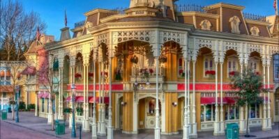 Early morning sunshine washes over the quaint and charming streetscape of a themed amusement park, highlighting the intricate detailing of its victorian-inspired architecture and vibrant floral arrangements. Disneyland Paris Emporium