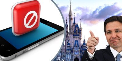 Ron DeSantis pointing with a smile while inside the Magic Kingdom Disney World park with a smartphone in the background.