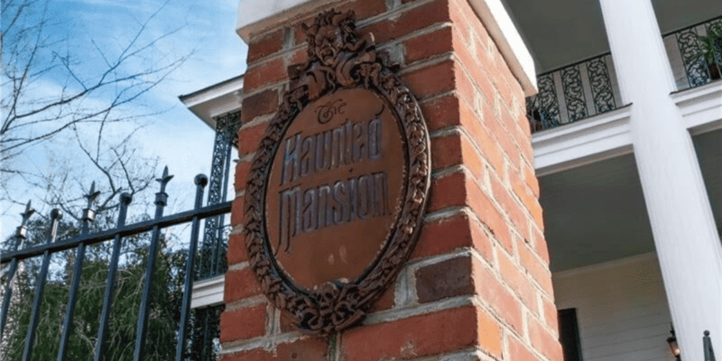 A brick pillar supports a circular metal sign reading "The Haunted Mansion" in ornate lettering. The pillar is part of a wrought iron fence, and in the background, a white, columned building is partially visible, hinting at the presence of the haunted mansion itself.