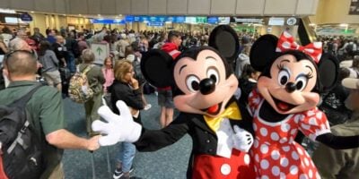 Mickey Mouse and Minnie Mouse greet and pose for a photo in a crowded Orlando International Airport terminal, surrounded by travelers and people with luggage as Disney guests get United Airlines ground stop.