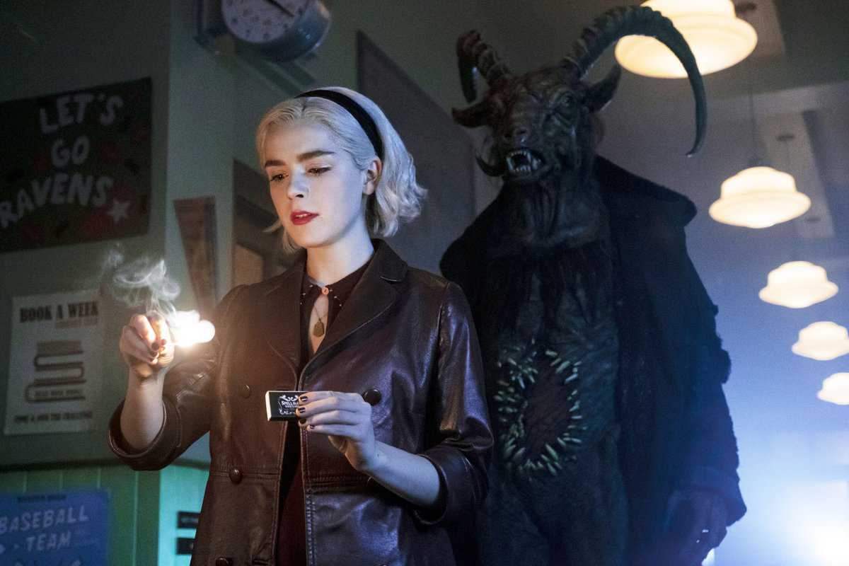 A woman with platinum blonde hair and a leather jacket gazes intently at the flame of a lighter, while an imposing creature with horns looms ominously in the background, under the eerie glow of the room's lighting.