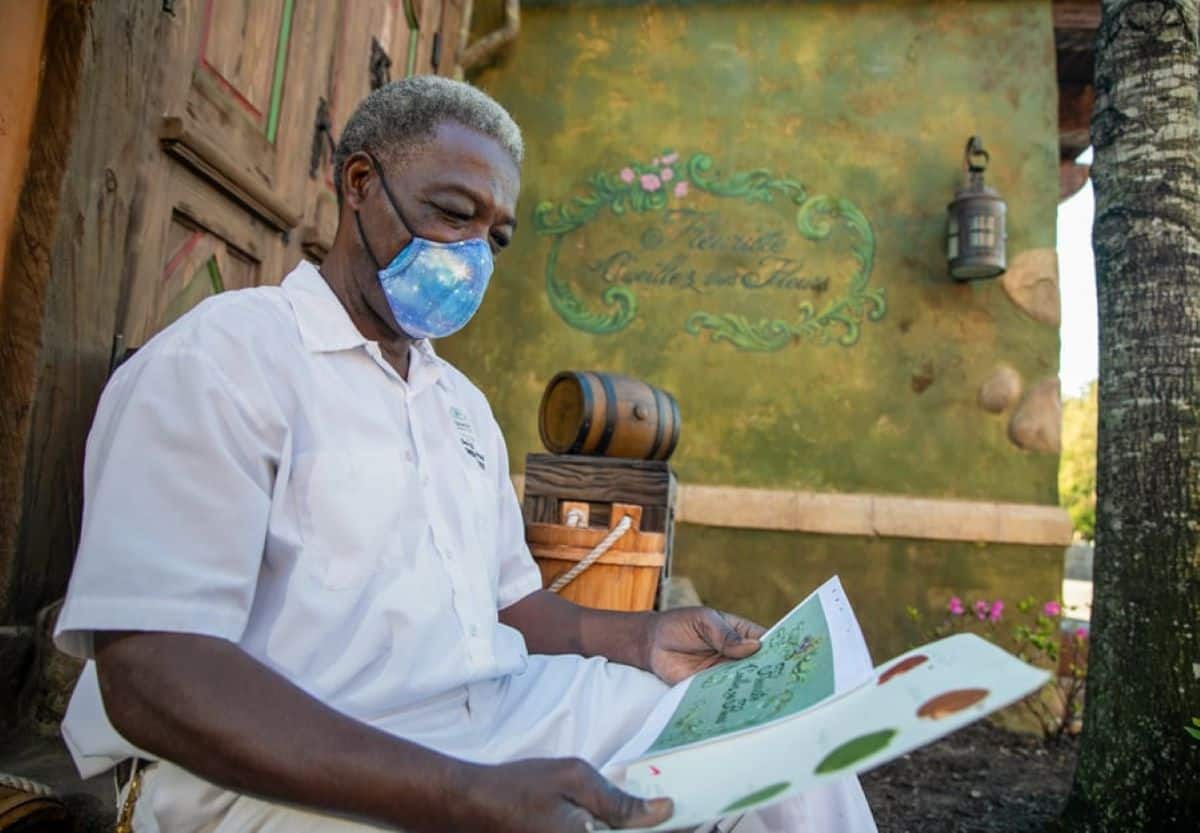 A man wearing a mask reviews a menu outside a charming eatery with a rustic ambiance in Disney World, captured on a sunny day.