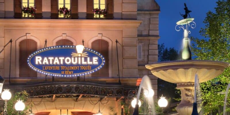 The image shows the entrance of "Ratatouille: L'Aventure Totalement Toquée de Rémy" at Disneyland Paris. The sign for the Ratatouille ride is brightly lit, and next to it is a decorative fountain with a figure on top. The scene is set during twilight, highlighted by soft illumination.