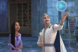 A man and a young girl in medieval attire marvel at a glowing orb he is levitating in a castle-like setting from Disney's Wish movie. The girl looks amazed and eager, while the man appears