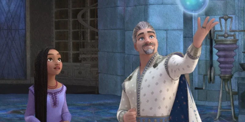 A man and a young girl in medieval attire marvel at a glowing orb he is levitating in a castle-like setting from Disney's Wish movie. The girl looks amazed and eager, while the man appears