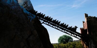 A Disney ride filled with people ascends a steep track against a backdrop of a clear blue sky, with rugged terrain and trees below.