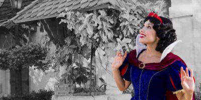 Woman dressed as snow white standing in front of a house with a black and white background, smiling and looking upward with one hand raised.