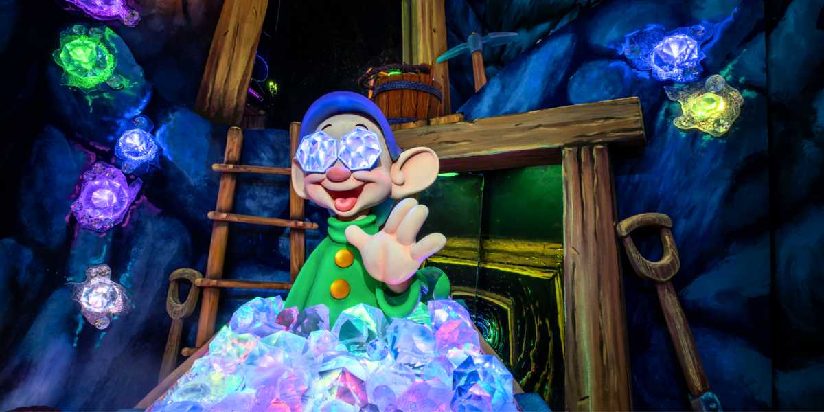 Dopey, a dwarf from snow white, smiling and waving in a colorful, jewel-lined mine shaft. he wears a green tunic and purple hat, and holds a shimmering, multi-colored crystal.