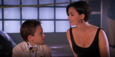 A young boy in a suit and an adult woman in a black dress smiling at each other in a warmly lit room with large windows in the background.