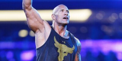 A muscular Dwayne Johnson in a tank top raising his right arm in a triumphant gesture, standing in a blue-lit arena. He has a tattoo on his left arm and is smiling confidently.