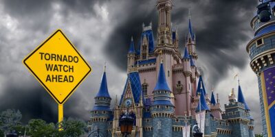 A dramatic image of the Disney Cinderella Castle under a dark, stormy sky with ominous clouds. in the foreground, a yellow road sign reads "tornado watch ahead.