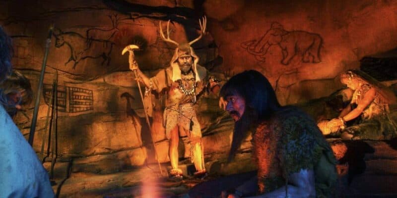 A scene depicting prehistoric humans in a cave, with one standing wearing animal skins and antlers while others sit by a fire, surrounded by cave paintings.
