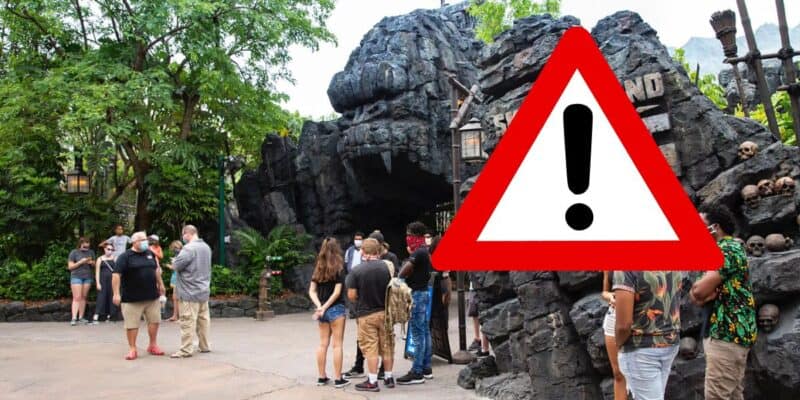 A warning sign with an exclamation mark overlays an image of a group of people standing near a rocky cave entrance at Universal Studios, suggesting caution or attention.