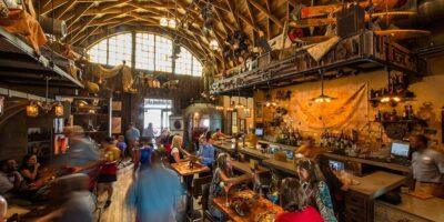 A bustling bar with patrons seated and standing, featuring eclectic decor with objects hanging from the ceiling, arched windows, and warmly lit surroundings.