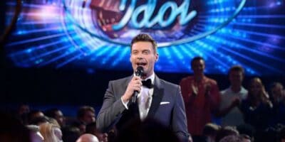 A charismatic host on stage at a talent show, energizing the audience with a microphone in hand, while the iconic "american idol" logo illuminates the background.