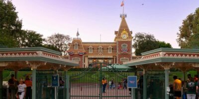 Entrance of disneyland with closed gates, featuring a building with a clock tower decorated with american flags, and people standing by the entrance.