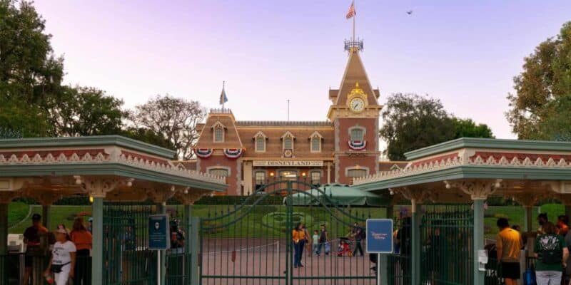 Entrance of disneyland with closed gates, featuring a building with a clock tower decorated with american flags, and people standing by the entrance.