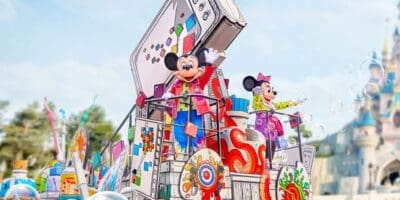 Mickey and Minnie mouse on a colorful parade float with an artistic backdrop of open books and vibrant designs, bubbles floating in the air, with a blurry castle in the background.